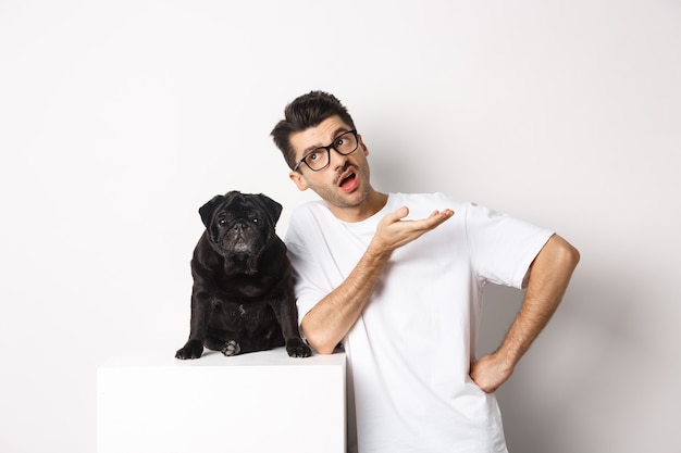 Image d'un gars hipster parlant avec son chien, l'air sceptique et gesticulant, ayant une conversation avec un mignon carlin noir, debout sur fond blanc.