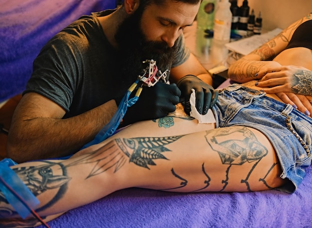 Image en gros plan de l'artiste tatoueur barbu fait un tatouage sur une jambe féminine.