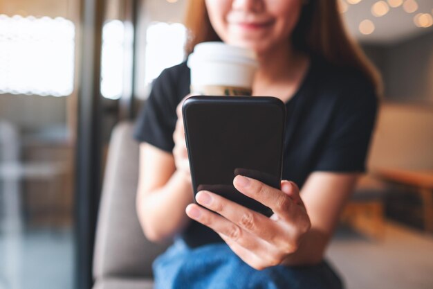 Image en gros plan d'une belle jeune femme asiatique tenant et utilisant un téléphone portable tout en buvant du café au café