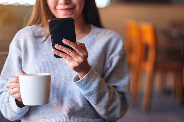 Image en gros plan d'une belle jeune femme asiatique tenant et utilisant un téléphone portable tout en buvant du café