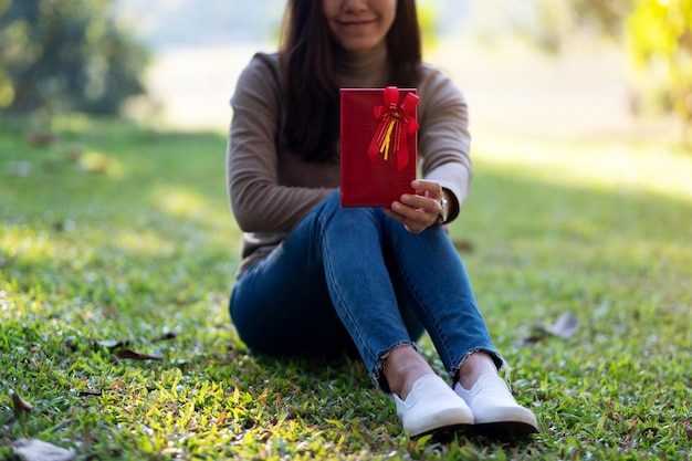 Image en gros plan d'une belle jeune femme tenant et donnant une boîte-cadeau rouge alors qu'elle était assise dans le parc