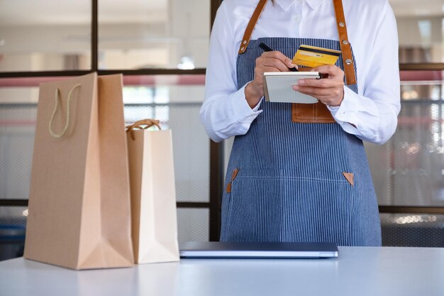 Image en gros plan d'une caissière tenant une carte de crédit tout en utilisant une tablette numérique et un ordinateur portable avec un sac à provisions sur le bureau de la caisse