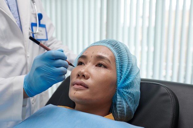 Image en gros plan d'un chirurgien plasticien laissant des marques sur le visage d'une jeune femme asiatique