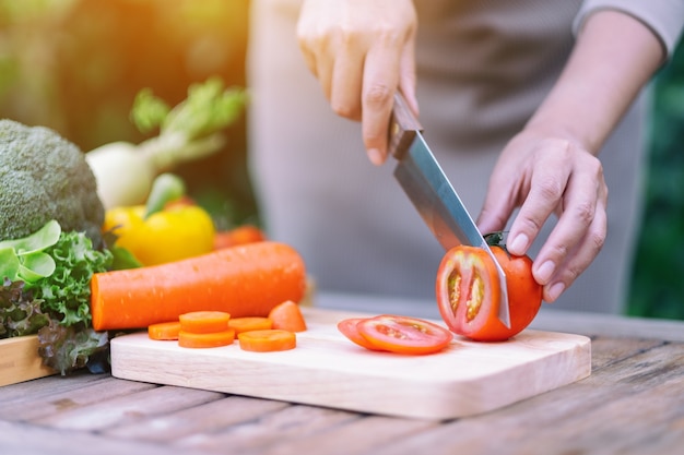 Image en gros plan d'une femme coupant et coupant la tomate au couteau sur une planche de bois