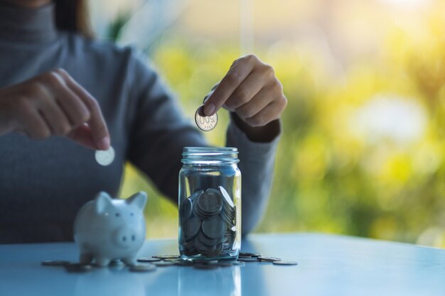 Image en gros plan d'une femme mettant une pièce dans une tirelire et un bocal en verre pour économiser de l'argent concept