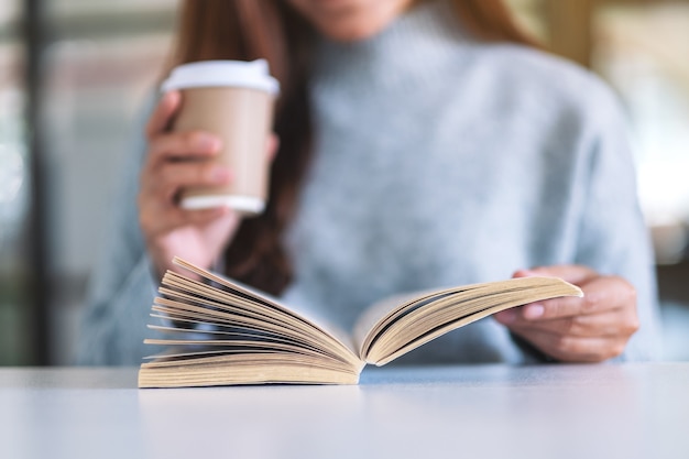 Image en gros plan d'une femme tenant et lisant un livre tout en buvant du café sur une table en bois