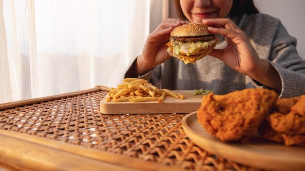 Image en gros plan d'une femme tenant et mangeant un hamburger et des frites avec du poulet frit sur la table à la maison
