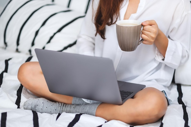 Image en gros plan d'une femme utilisant et travaillant sur un ordinateur portable, buvant du café assis sur un lit confortable blanc à la maison