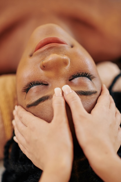 Image en gros plan d'une jeune femme afro-américaine recevant un massage professionnel de la tête et du visage au salon spa