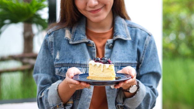 Image en gros plan d'une jeune femme asiatique tenant et montrant une assiette de gâteau au fromage aux bleuets