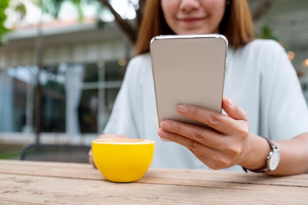 Image en gros plan d'une jeune femme asiatique tenant et utilisant un téléphone portable tout en buvant du café