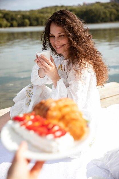 Photo image en gros plan d'une jeune femme souriante aux longs cheveux bouclés vêtue d'une robe blanche servant une tasse de thé et un croissant à l'extérieur