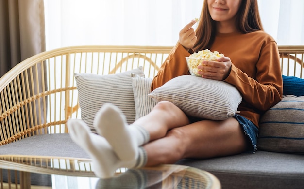 Image en gros plan d'une jeune femme tenant et mangeant du maïs soufflé en regardant la télévision et assise sur un canapé à la maison