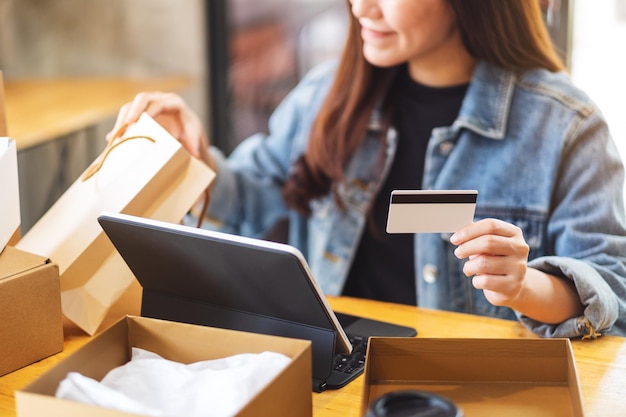 Image en gros plan d'une jeune femme utilisant une tablette et une carte de crédit pour faire des achats en ligne, ouvrant des sacs à provisions et une boîte à colis postal sur la table
