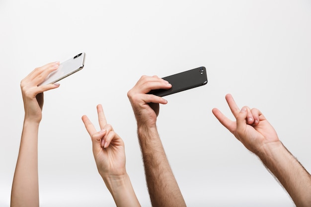 L'image en gros plan des mains d'un homme et d'une femme isolées sur un mur blanc tenant des téléphones portables prend un selfie montrant un geste de paix.