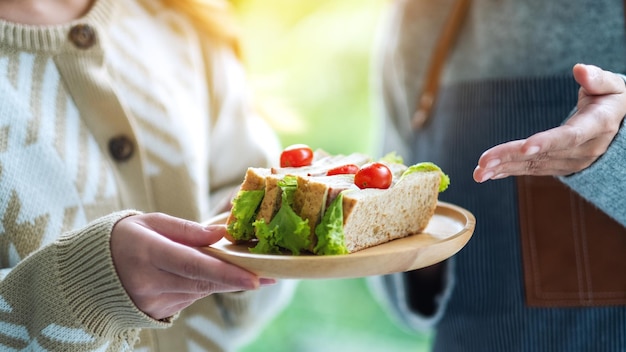 Image en gros plan d'une serveuse tenant et servant un sandwich au blé entier dans une assiette en bois au client