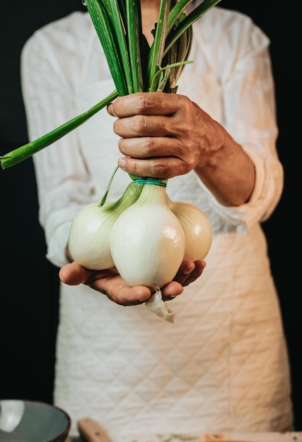 Image en gros plan d'une vieille femme chef de cuisine attrapant des oignons et montrant à la caméra avant de les préparer dans une cuisine saine Cuisine rustique préparée par le chef Préparation d'un ingrédient pour un repas bio