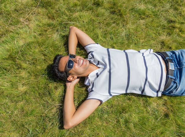 Image d'en haut du jeune garçon aux cheveux noirs allongé au soleil sur l'herbe avec des lunettes de soleil noires, une chemise blanche et un jean