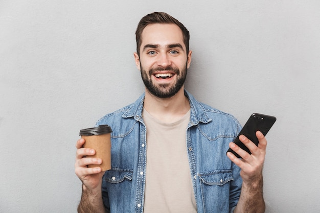 Image d'heureux homme européen ayant barbe à l'aide de smartphone et de boire du café à emporter isolé sur mur gris