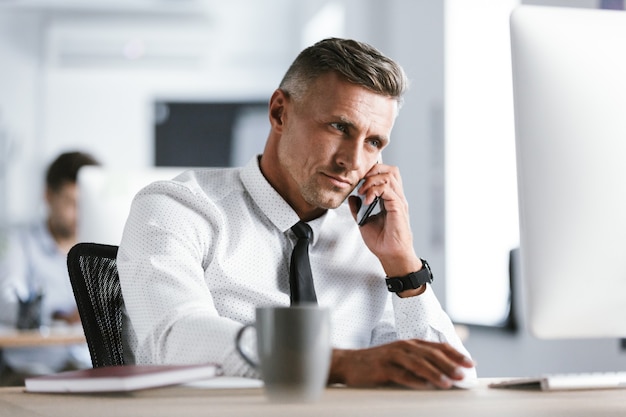 Image d'homme d'affaires adulte de 30 ans portant chemise blanche et cravate assis au bureau au bureau par ordinateur, et parler sur smartphone