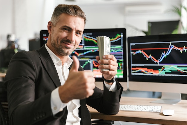 Image d'un homme d'affaires attrayant de 30 ans portant un costume tenant un paquet d'argent tout en travaillant au bureau avec des graphiques et des tableaux sur ordinateur