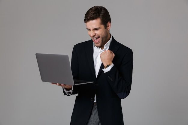 Image d'homme d'affaires extatique de 30 ans en costume se réjouissant tout en utilisant un ordinateur portable, isolé sur mur gris