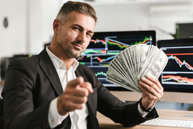 Image d'homme d'affaires heureux 30 s portant costume tenant un ventilateur d'argent tout en travaillant au bureau avec des graphiques et des tableaux sur ordinateur