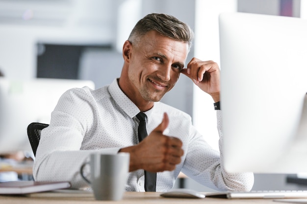 Image d'homme d'affaires joyeux 30 s portant chemise blanche et cravate assis au bureau au bureau par ordinateur, et montrant le pouce vers le haut