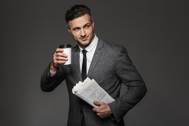 Image de l'homme d'affaires occupé habillé en costume d'affaires à côté et de boire du café à emporter avec du papier journal à la main, isolé sur mur gris