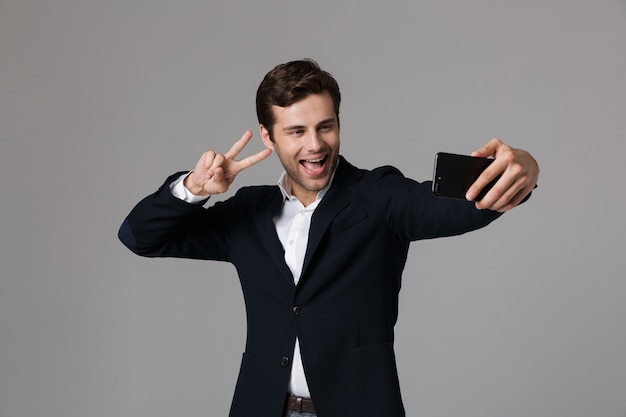 Image d'homme d'affaires optimiste des années 30 en costume officiel montrant le signe de la paix tout en prenant selfie photo sur téléphone portable, isolé sur mur gris
