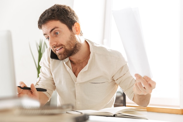 Image d'homme d'affaires sérieux de 30 ans portant une chemise blanche, parler au téléphone mobile, alors qu'il était assis à table dans un bureau moderne