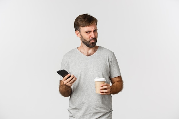 Image d'homme barbu confus debout avec café et téléphone portable, à la perplexité