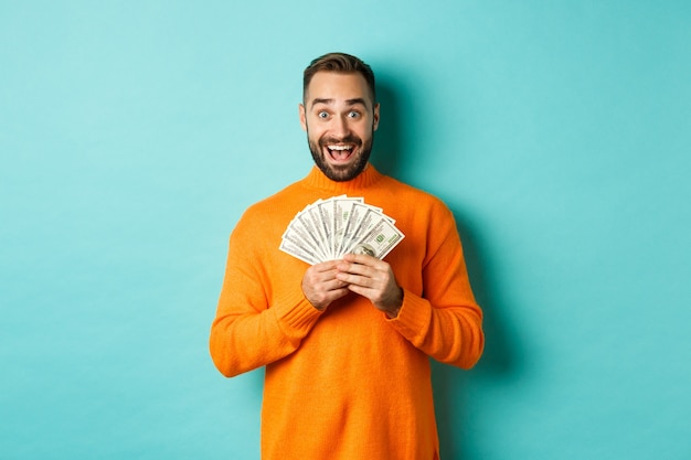 Image d'un homme barbu excité tenant de l'argent et se réjouissant, remportant un prix en espèces, debout sur fond bleu clair