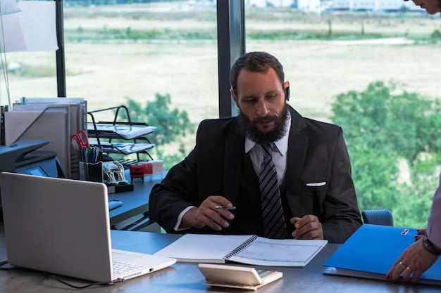 Image d'un homme barbu portant une cravate et un costume noir assis au bureau à table, tenant un stylo à la main. Homme avec des documents et des dossiers travaillant avec un ordinateur portable au bureau au bureau. Document à la recherche d'un avocat