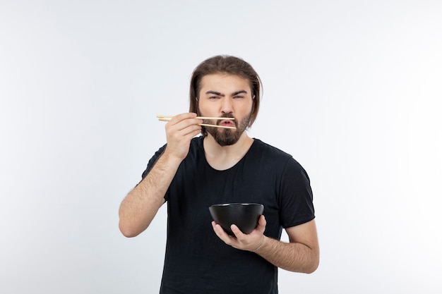 Image d'homme barbu tenant un bol avec des baguettes sur un mur blanc.