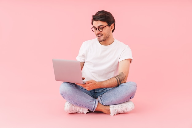 Image d'un homme brune à lunettes à l'aide d'un ordinateur portable assis avec les jambes croisées isolées