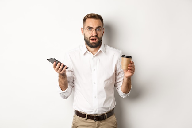 Image d'un homme buvant du café, se sentant confus au sujet d'un message étrange sur un téléphone mobile, debout sur fond blanc.