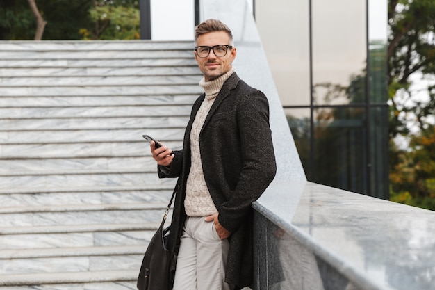 Image d'un homme caucasien de 30 ans portant des lunettes, marchant dans la rue de la ville et à l'aide de smartphone