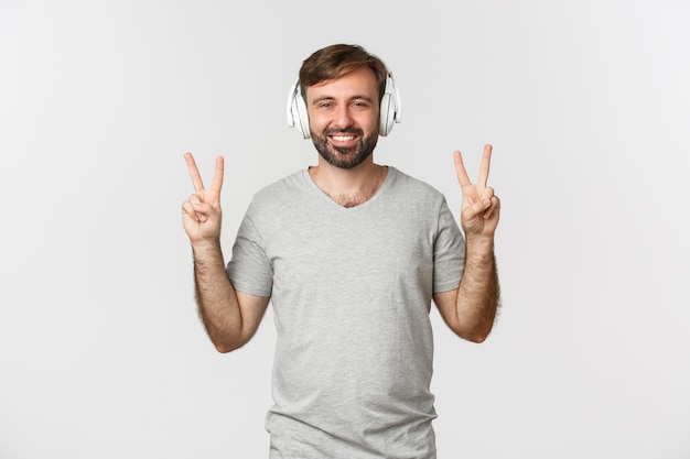 Image d'un homme caucasien heureux en t-shirt gris, écouter de la musique dans des écouteurs et montrer des signes de paix