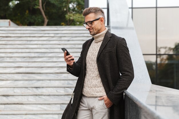 Image d'un homme européen de 30 ans portant des lunettes, marchant dans la rue de la ville et à l'aide de smartphone