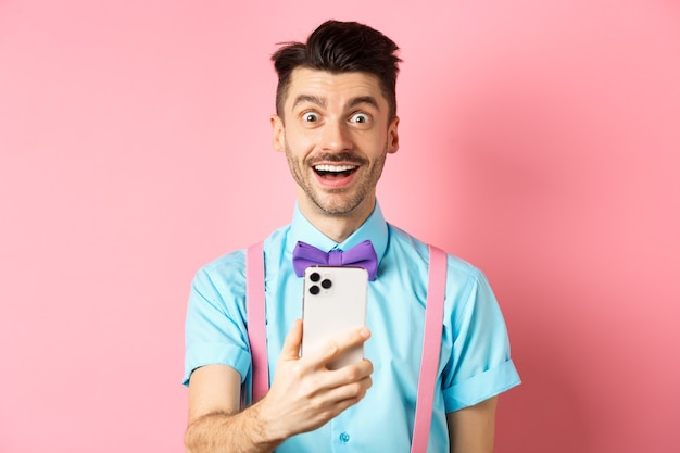 Image d'un homme excité vérifiant l'offre promotionnelle en ligne, tenant un smartphone et regardant la caméra surpris, debout heureux sur le rose.