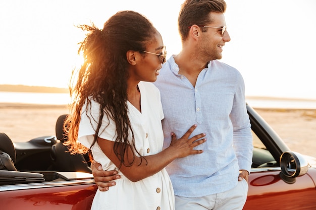 Image d'un homme et d'une femme heureux couple multiethnique souriant et s'embrassant ensemble en se tenant debout en voiture à l'extérieur