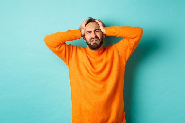 Image d'un homme frustré en pull orange, tenant les mains sur la tête et se sentant en détresse, debout anxieux