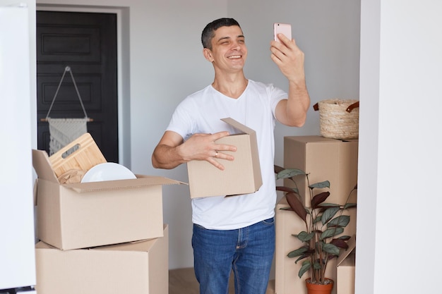Image d'un homme heureux souriant portant un T-shirt blanc et un jean debout avec une boîte en carton, tenant un smartphone et prenant un selfie dans un nouvel appartement ou diffusant en direct.