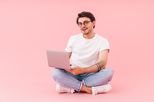 Image d'un homme joyeux à lunettes utilisant un ordinateur portable assis avec les jambes croisées isolées