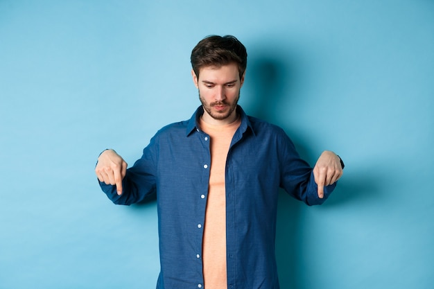 Image d'un homme moderne dans des vêtements décontractés, regardant et pointant du doigt vers le bas sur un espace vide, lisant le logo, debout sur fond bleu.