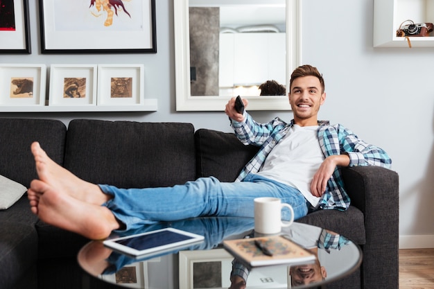 L'image d'un homme à poils joyeux vêtu d'une chemise dans une impression de cage se trouve sur un canapé à la maison et tient la télécommande tout en regardant la télévision.