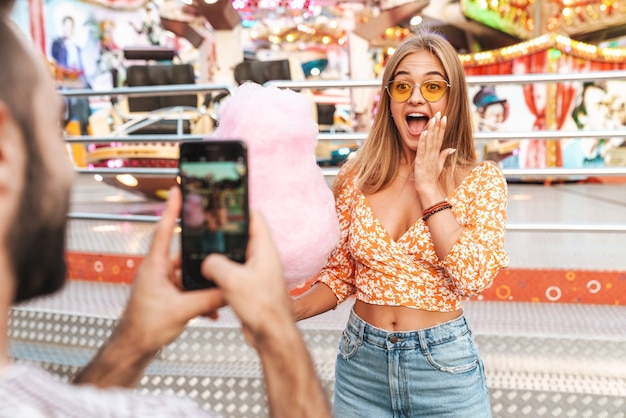 Image d'un homme prend une photo de sa femme émotionnelle surprise marchant à l'extérieur dans un parc d'attractions par téléphone portable.