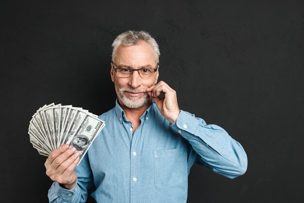 Image d'un homme riche d'âge moyen des années 60 aux cheveux gris tenant de l'argent fan de 100 dollars et touchant sa moustache grise, isolé sur mur noir