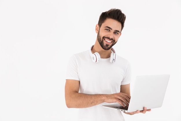 Image d'un homme satisfait non rasé avec des écouteurs sur le cou souriant et utilisant un ordinateur portable isolé sur un mur blanc